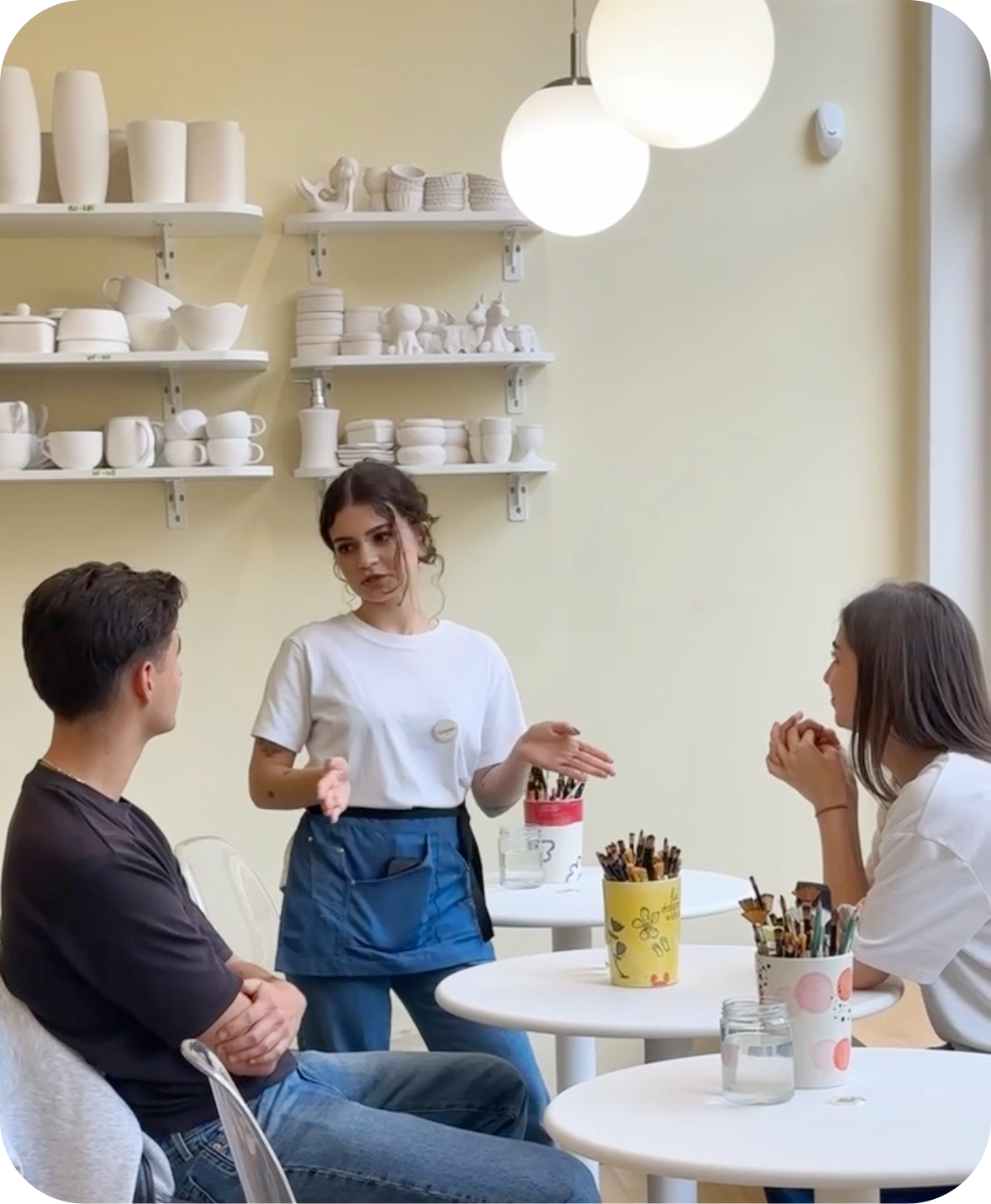 A café employee explains to a couple how to paint ceramics at white tables with shelves of white mugs behind them at Create Cafe in Warsaw