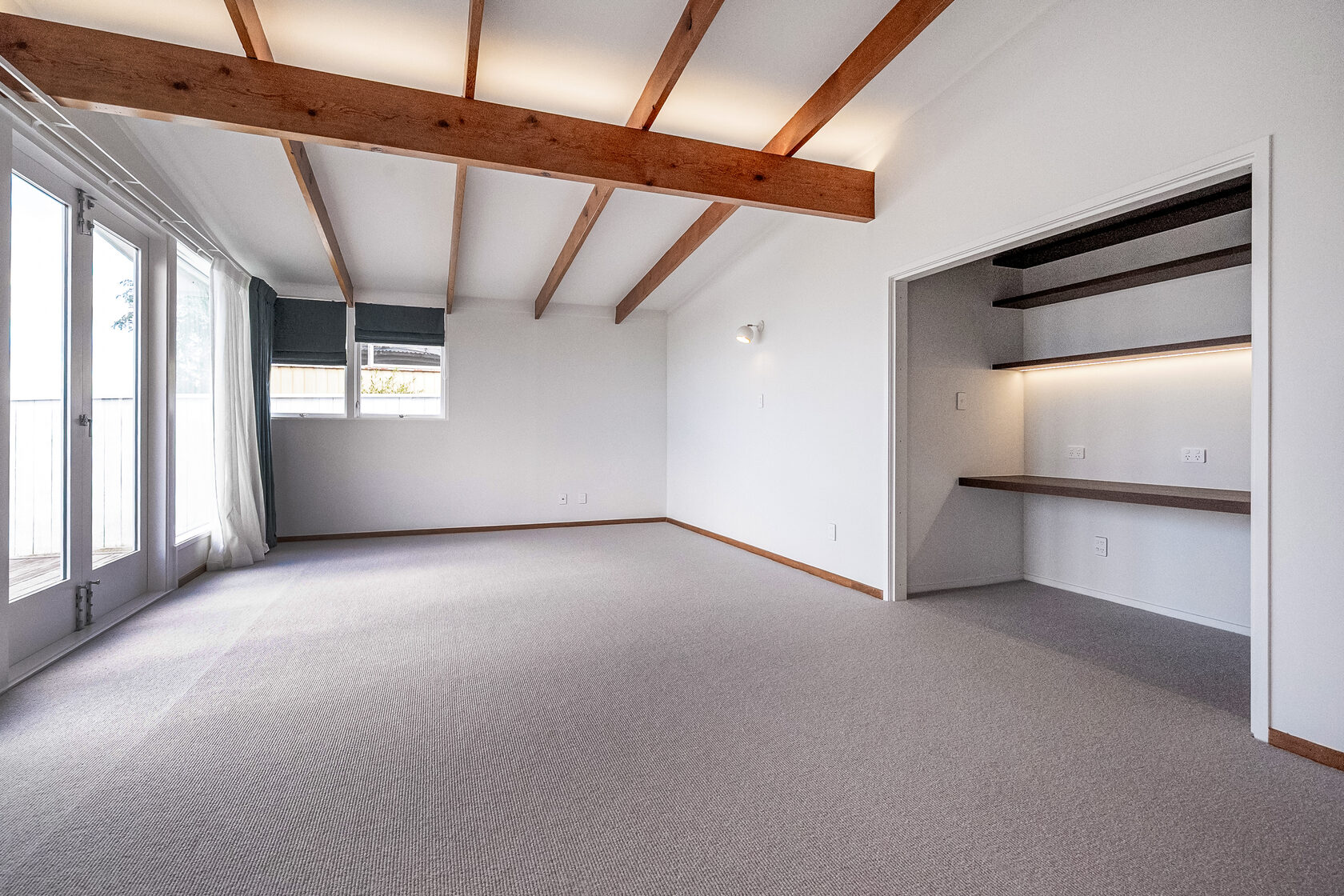 Bright living room with exposed timber ceiling beams, built-in shelving nook with LED lighting, and large windows with curtains.