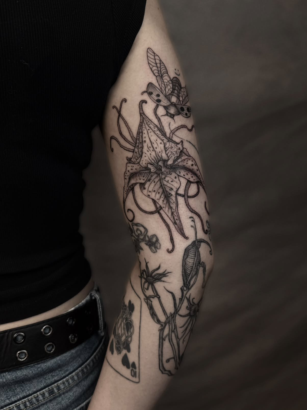 Upper-arm tattoo of a drooping flower with speckled pointed petals, long tendrils and a beetle with open wings above it.