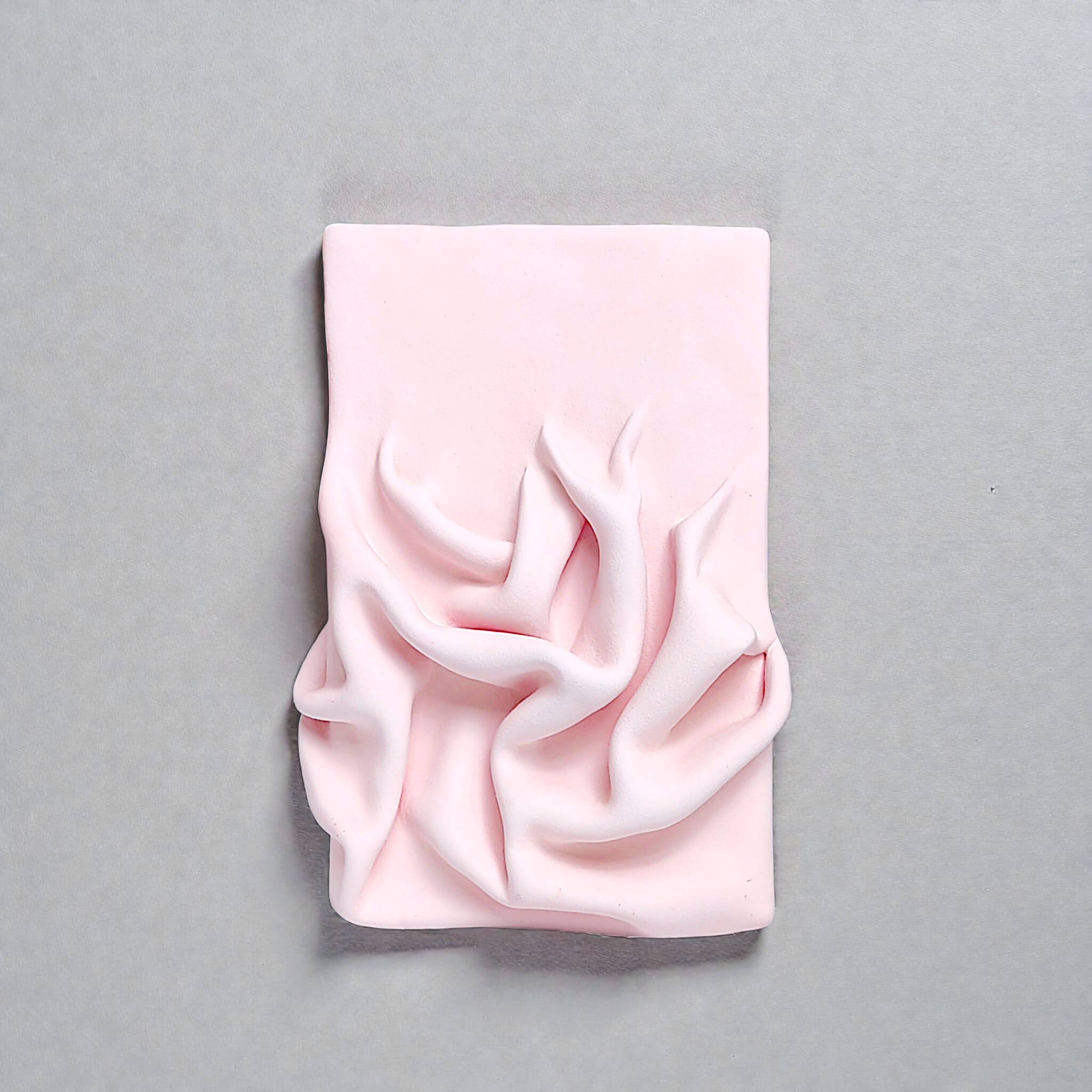 Handmade pastel pink ceramic wall art with sculptural, fabric-like folds. Modern textured artwork for interior decor in a minimal or soft-toned space.