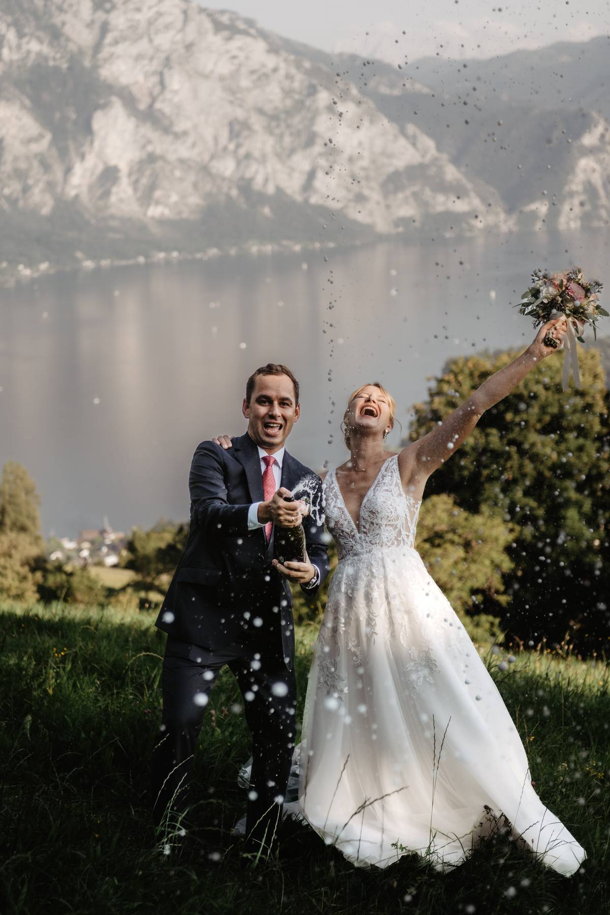 Mountain wedding couple popping champagne outdoors, joyful spray with lake and peaks in the background