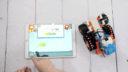 A LEGO&reg; vehicle on tracks driving across a white table, next to a tablet