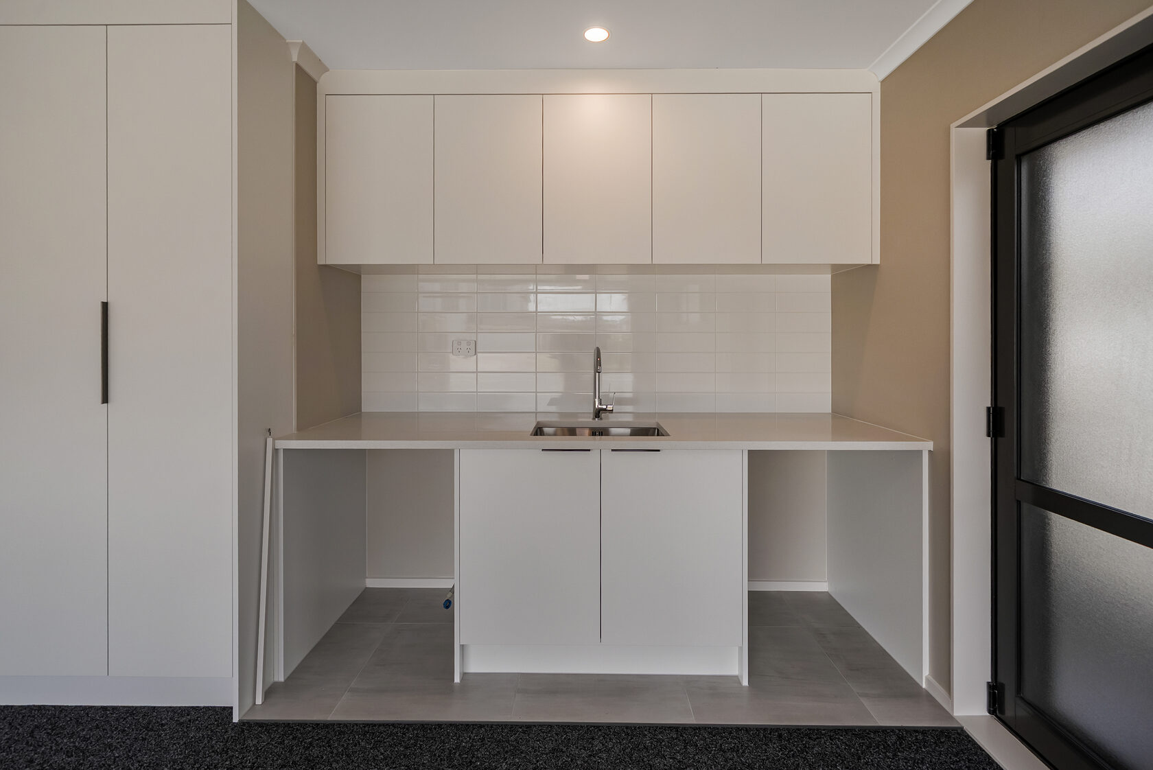 Flat Bush home renovation: Minimalist laundry area with white cabinetry, tiled splashback, stainless steel sink, and neutral finishes.