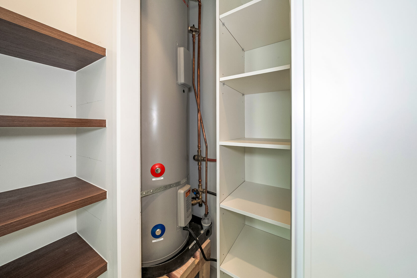 Utility cupboard with hot water cylinder, exposed plumbing, and built-in storage shelves.